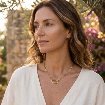 A woman with wavy brown hair wearing a white V-neck top and a delicate gold chain necklace with a small green rectangular accent, photographed outdoors with blurred foliage and stone in the background.