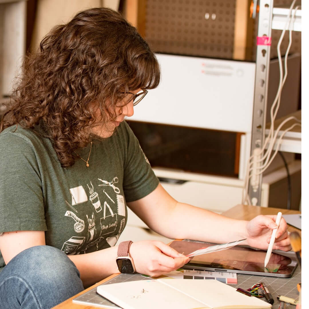 A woman sitting at a desk working on a tablet while holding a color swatch