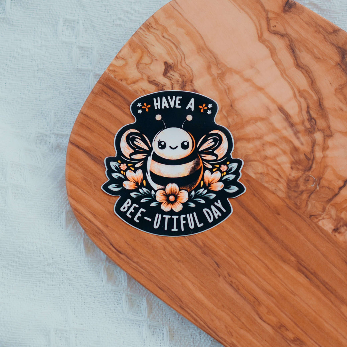 Have a Bee-U-tiful Day sticker with a cheerful bee design and flowers on a wooden surface.