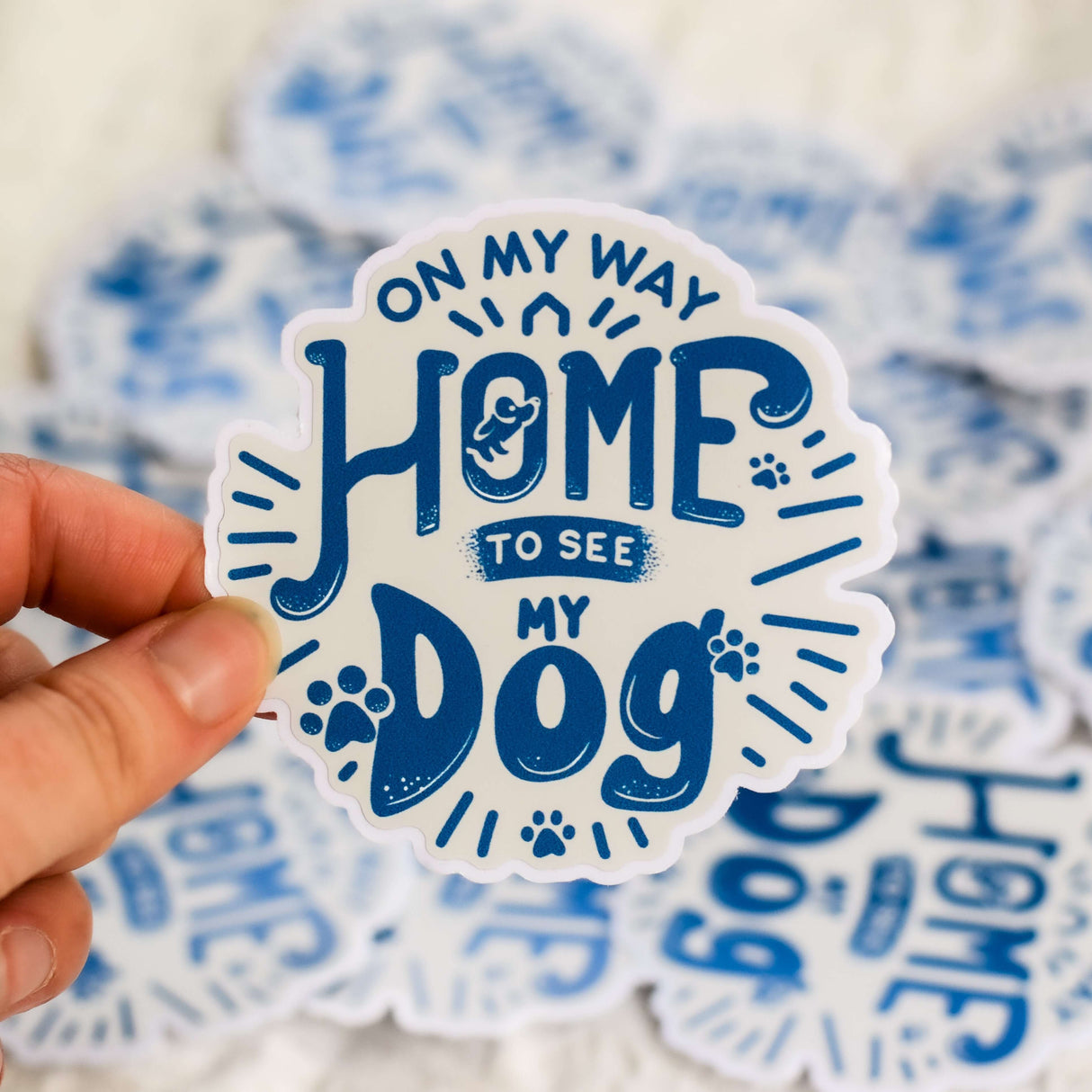 Person holding "On My Way Home to See My Dog" sticker with blue text and paw print design