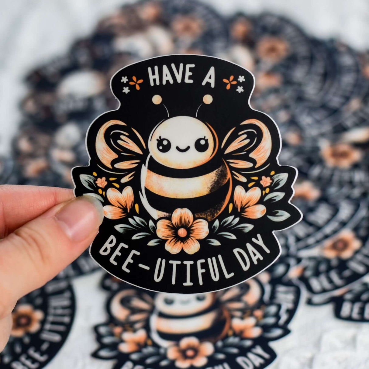 Hand holding a Have a Bee-U-tiful Day sticker featuring an adorable bee and flowers, made from high-quality, weather-resistant vinyl.