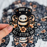 Hand holding a Have a Bee-U-tiful Day sticker featuring an adorable bee and flowers, made from high-quality, weather-resistant vinyl.