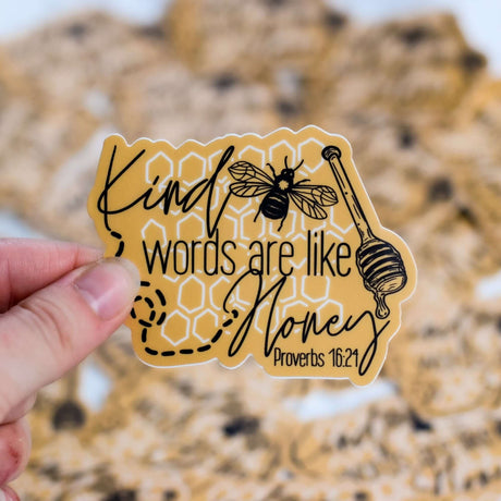 Hand holding "Kind Words Are Like Honey" vinyl sticker with Proverbs 16:24 verse and bee illustration.