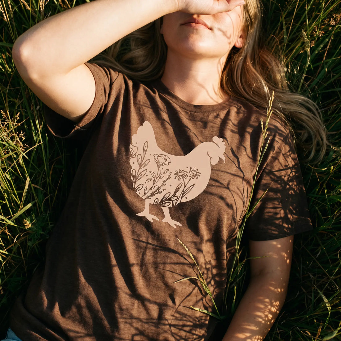 A woman lying in grass outdoors wearing a brown short-sleeve T-shirt with a large pale peach chicken graphic filled with floral details on the front.