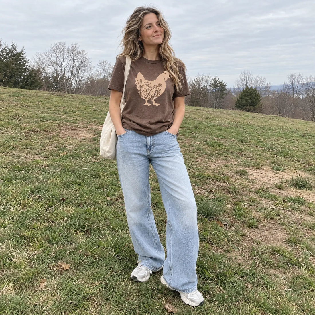 A woman stands on a grassy hill wearing a brown graphic t-shirt with a chicken design, light-wash wide-leg jeans, and white sneakers, carrying a cream canvas tote bag.