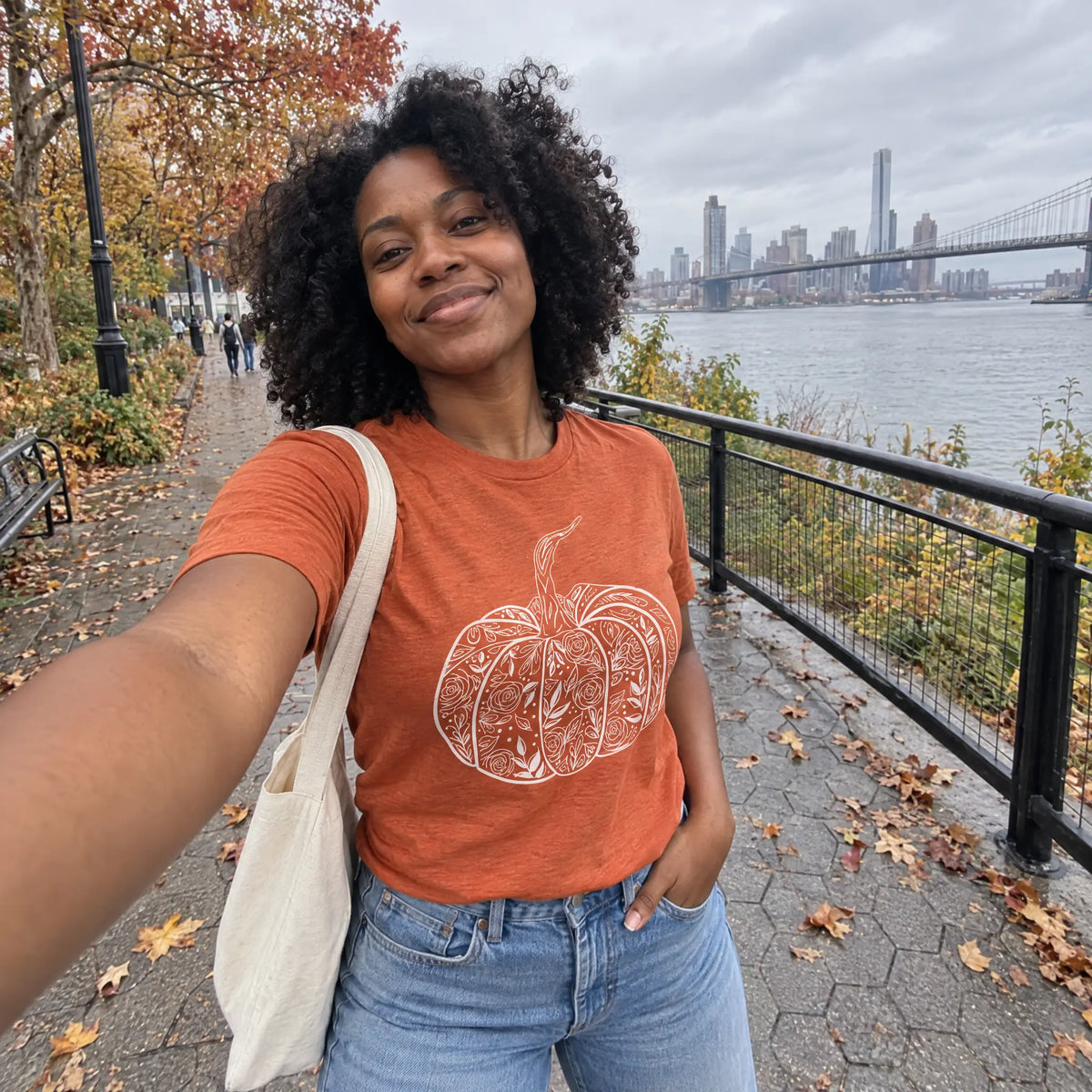 Smiling woman taking a selfie in a heather autumn orange floral pumpkin graphic tee on a riverside walkway