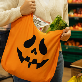 Person holding an orange tote bag with a pumpkin face design, filled with groceries, in a store setting.
