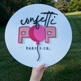 Hand holding custom round sign with "Confetti Pop Party Co" text, ideal for branding, events, and home decor