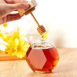 Engraved hexagon glass honey dispenser with dipper, featuring wooden lid, filled with honey on a wooden table with flowers in background.