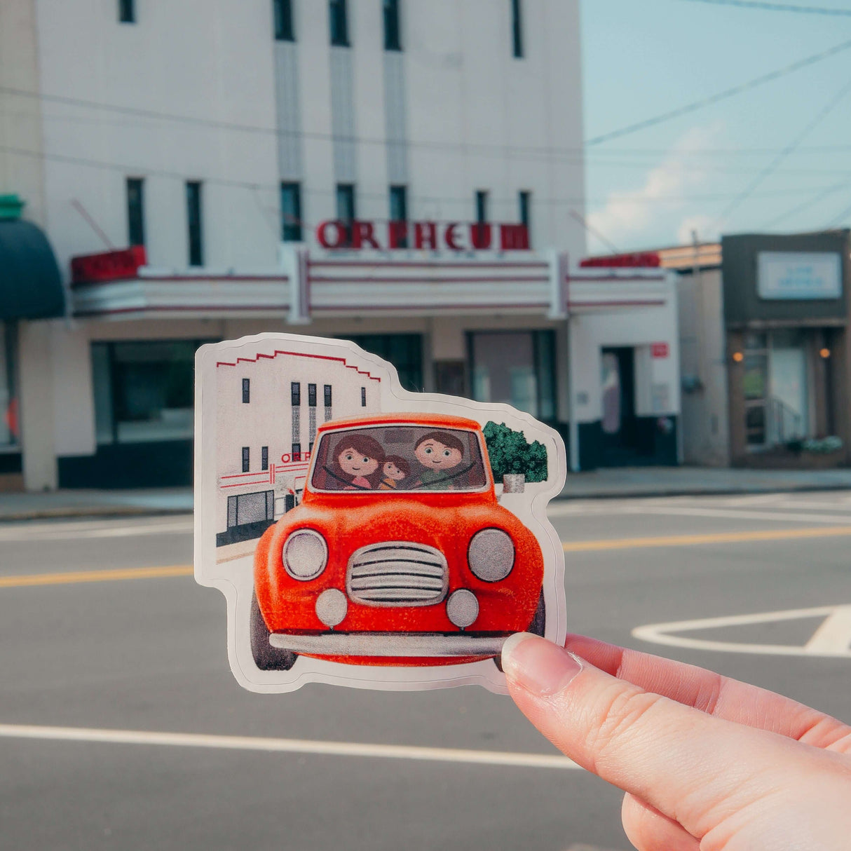 Person holding Max's Festival Adventure sticker with kids in a red car in front of the Orpheum theater