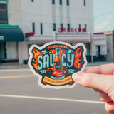 Hand holding NC Hot Sauce Contest & Festival sticker in front of historic building in North Carolina.