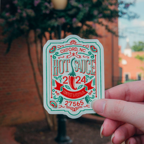 "Hand holding NC Hot Sauce Contest & Festival sticker with colorful design, background of outdoor setting in Oxford, North Carolina"