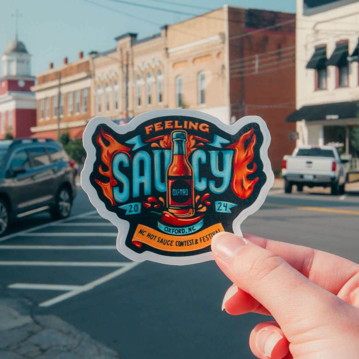 "Hand holding NC Hot Sauce Contest & Festival sticker with 'Feeling Saucy' design in downtown Oxford, NC"