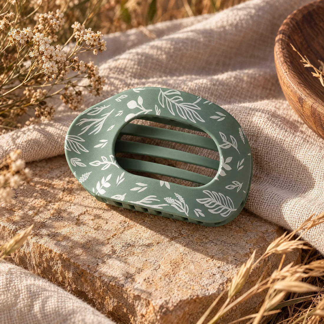 Sage green claw hair clip with a matte finish and a white hand-drawn botanical print of leaves, branches, and berries. The oval clip has an open cutout top and interlocking teeth. It rests on a sunlit stone slab draped with natural linen, next to dried wildflowers, golden grasses, and a wooden bowl.