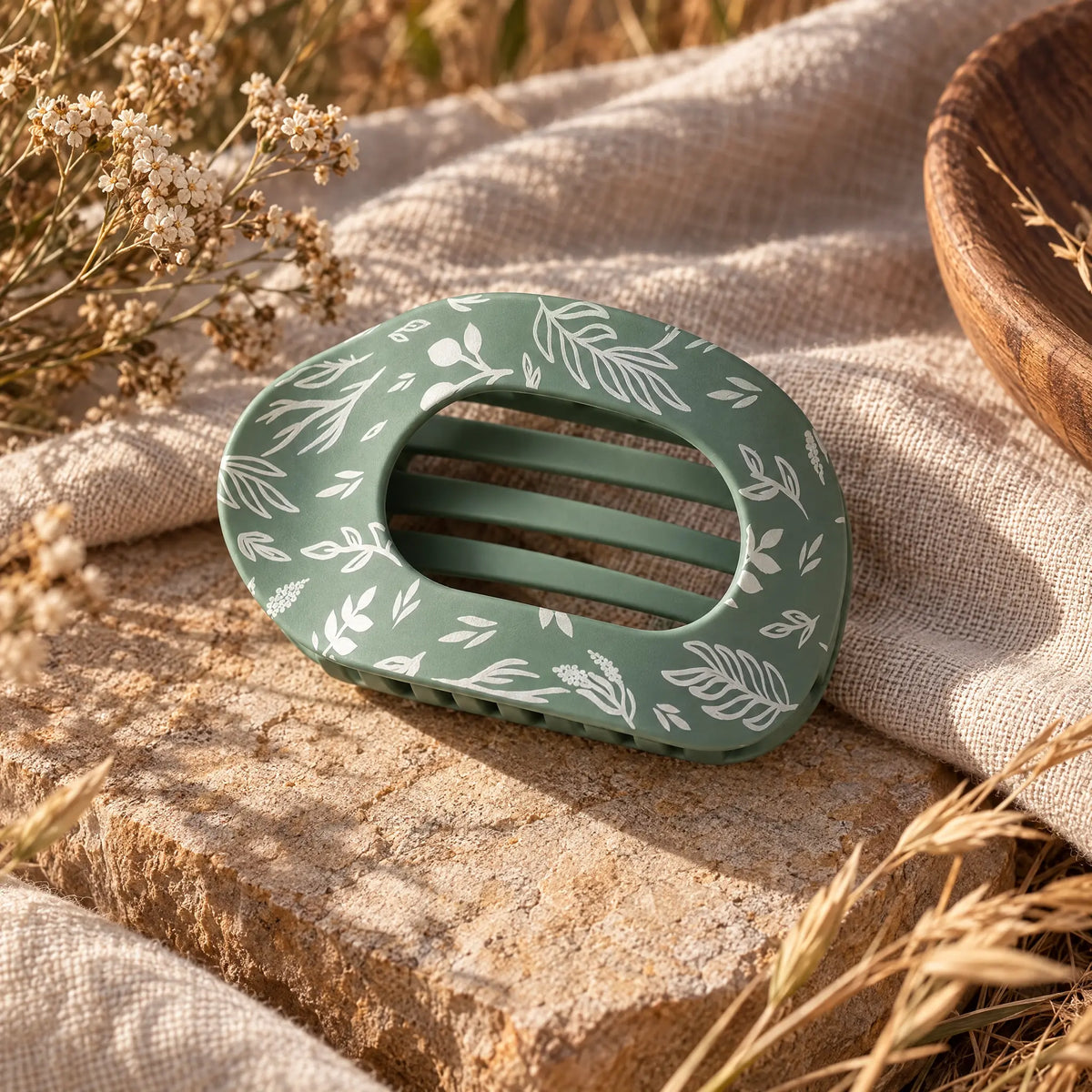 Sage green claw hair clip with a matte finish and a white hand-drawn botanical print of leaves, branches, and berries. The oval clip has an open cutout top and interlocking teeth. It rests on a sunlit stone slab draped with natural linen, next to dried wildflowers, golden grasses, and a wooden bowl.
