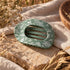 Sage green claw hair clip with a matte finish and a white hand-drawn botanical print of leaves, branches, and berries. The oval clip has an open cutout top and interlocking teeth. It rests on a sunlit stone slab draped with natural linen, next to dried wildflowers, golden grasses, and a wooden bowl.