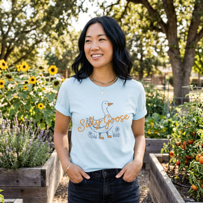 A woman wearing a light blue t-shirt with a graphic of a goose and the text "Silly Goose" stands in a garden surrounded by flowers and plants.