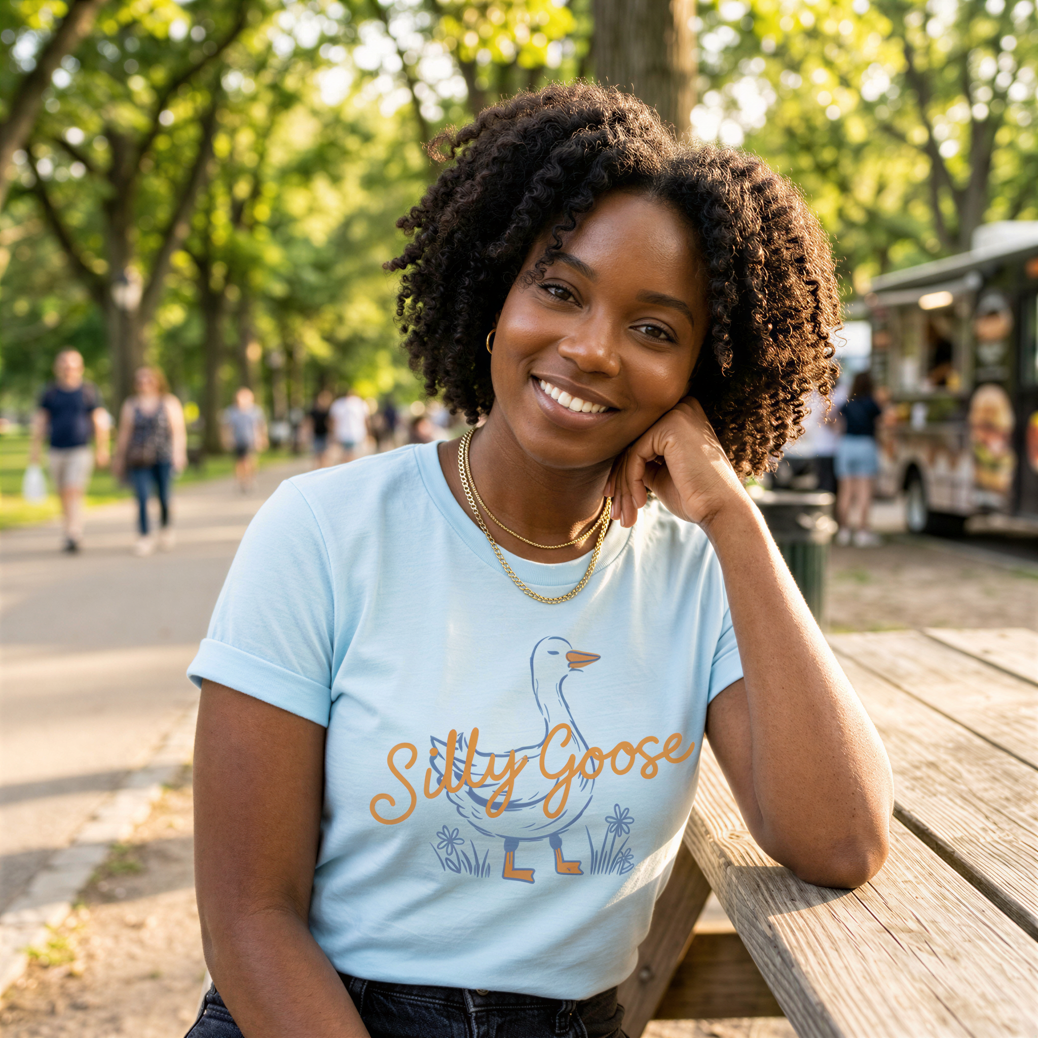 A smiling woman with curly hair wearing a light blue t-shirt with a graphic of a goose and the text "Silly Goose" is sitting on a wooden bench in a park.