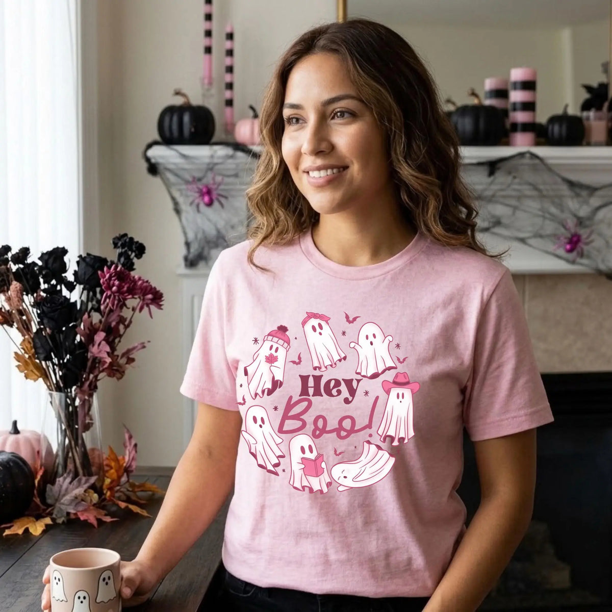 Smiling woman in a heather pink t-shirt with a &amp;quot;Hey Boo!&amp;quot; Halloween graphic. Cute white ghosts circle the words, some wearing a beanie, a bow, or a pink cowboy hat, and one is reading a book. She holds a ghost print mug in a living room decorated for Halloween. The mantel behind her has black pumpkins, pink and black striped candles, and spider webs, and a vase of dark dried flowers sits beside her.