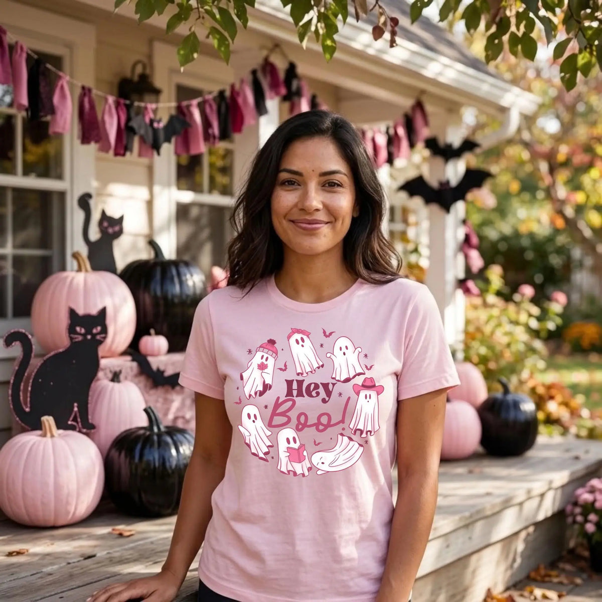 Smiling woman in a light pink t-shirt with a &amp;quot;Hey Boo!&amp;quot; Halloween graphic. Cute white ghosts circle the words, some wearing a beanie, a bow, or a pink cowboy hat, and one is reading a book. She stands on a porch decorated for Halloween with pink and black pumpkins, black cat cutouts, paper bats, and a pink and black fabric garland.