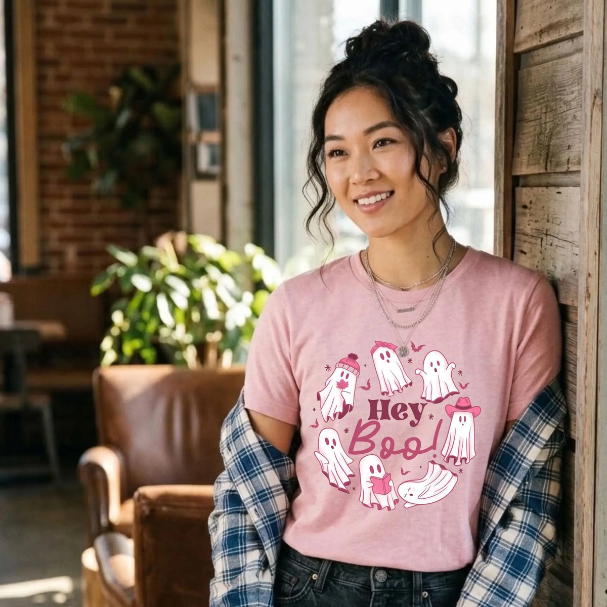 Smiling woman in a heather pink t-shirt with a &amp;quot;Hey Boo!&amp;quot; Halloween graphic. Cute white ghosts circle the words, some wearing a beanie, a bow, or a pink cowboy hat, and one is reading a book. She wears it with a blue plaid flannel slipping off her shoulders, black jeans, and layered necklaces. She leans against a rustic wood wall in a sunny cafe with a leather armchair.