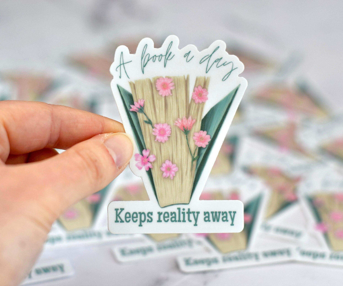 Vinyl sticker from Winks Design Studio featuring the quote "A Book a Day Keeps Reality Away" with a book illustration.