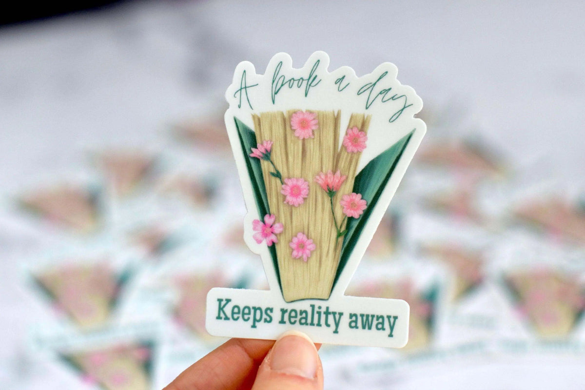 Vinyl sticker featuring the quote "A Book a Day Keeps Reality Away" by Winks Design Studio, LLC, with book and whimsical design elements.