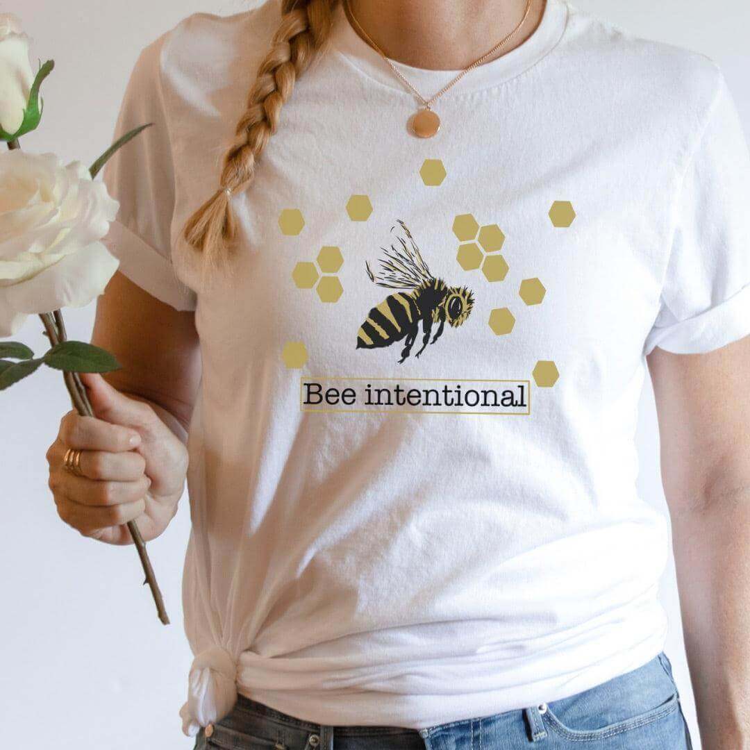 Woman wearing SCBA Bee Intentional T-Shirt with bee and hexagon design, holding a white rose